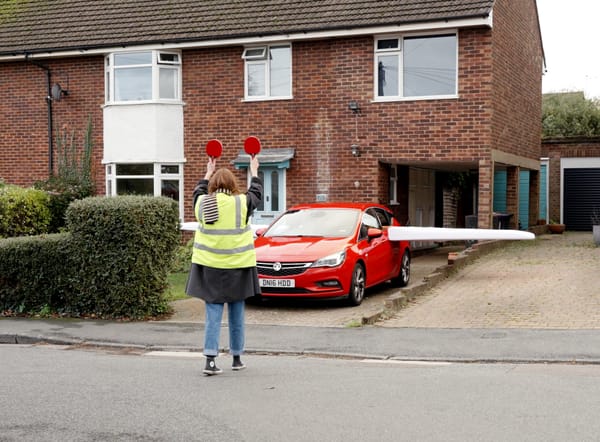 An aircraft marshall guides a car with wings taxi on the runway in front of a suburban house